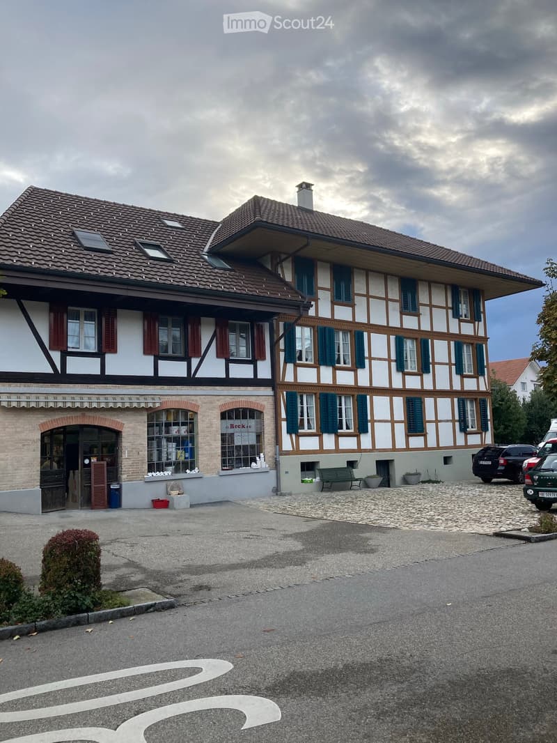Two-story traditional building with white and brown timber framing, multiple windows with shutters, and a tiled roof. A small entrance area with a bench and potted plants is visible.