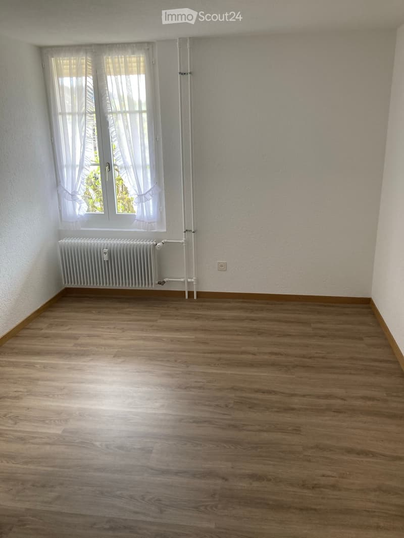 Empty room with wooden flooring, white walls, a window with white curtains, and a radiator below the window.