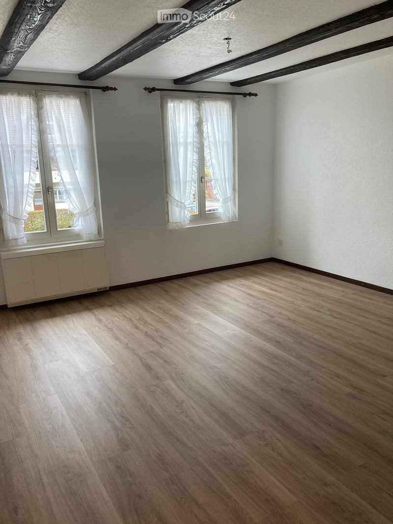 Empty room with wooden flooring, white walls, and large windows with white curtains. Exposed wooden beams on the ceiling.