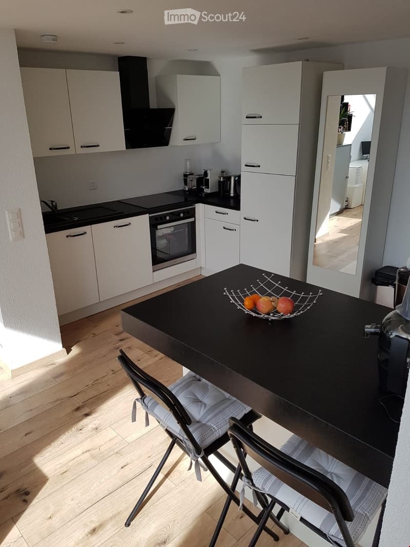 Modern kitchen with white cabinets, black countertops, and stainless steel appliances. Includes a coffee maker, fruit bowl, and a small dining area with a table and chairs.