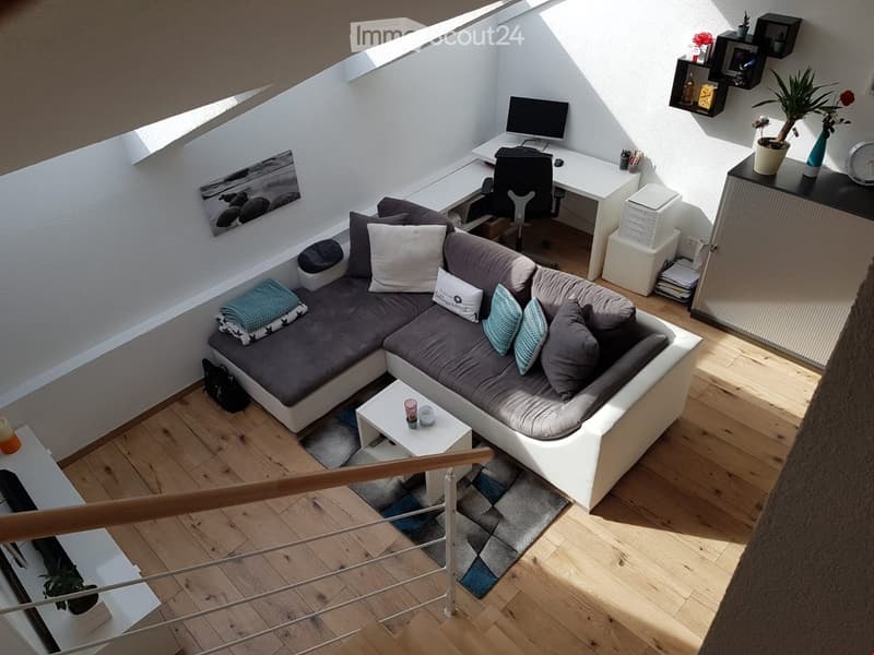 A modern, multi-functional living space with a grey sectional sofa, a small coffee table, a desk with a computer, and a potted plant. The room features wooden flooring and a staircase leading to another level.