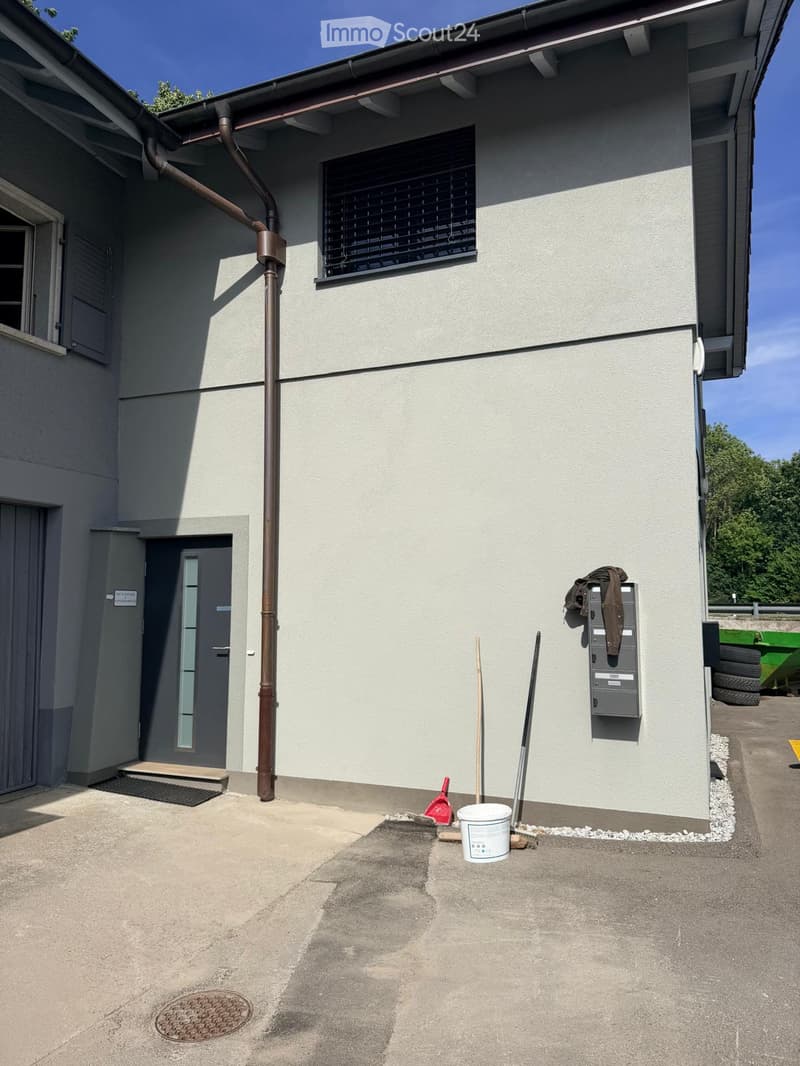 Exterior view of a two-story building with a light gray facade, a window with blinds, a door, and a utility box on the wall. Cleaning tools and a bucket are placed on the ground near the entrance.