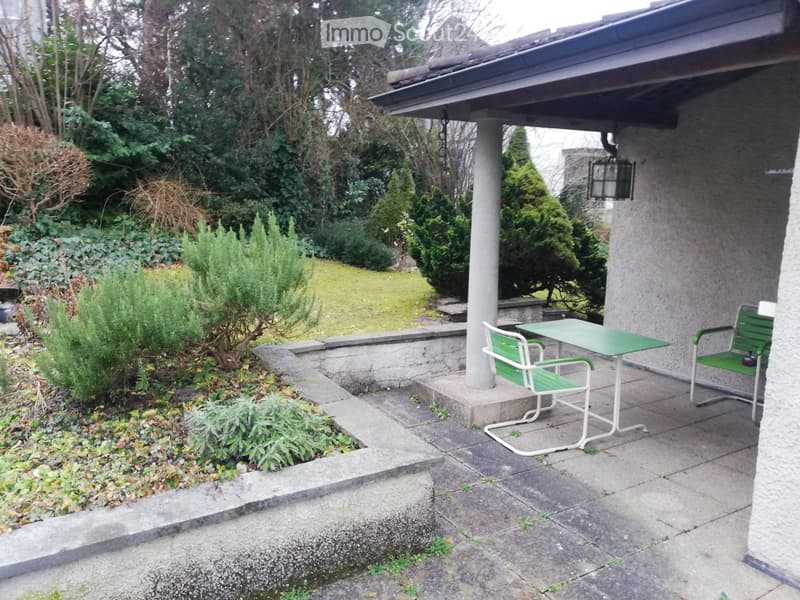 A well-maintained garden with a patio area featuring a green metal table and two matching chairs, surrounded by neatly trimmed bushes and trees.