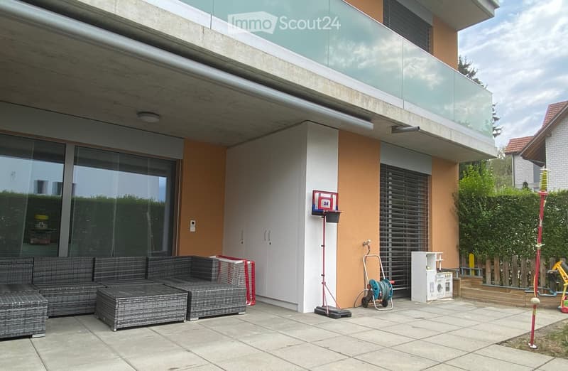 Modern outdoor patio area with a grey wicker sofa and table, a washing machine, and a hose reel, adjacent to a contemporary house with large glass windows and a balcony.
