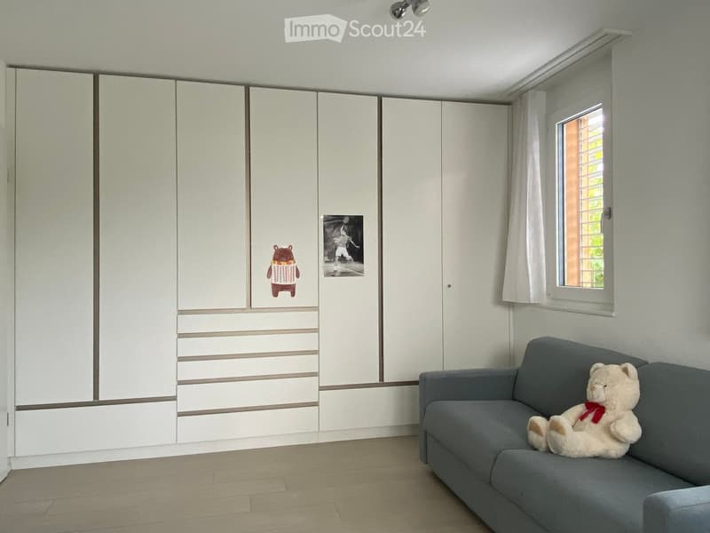 A furnished room with a grey sofa, a large white wardrobe, and a teddy bear toy on the sofa. The room has a window with white curtains and a small photo on the wardrobe.