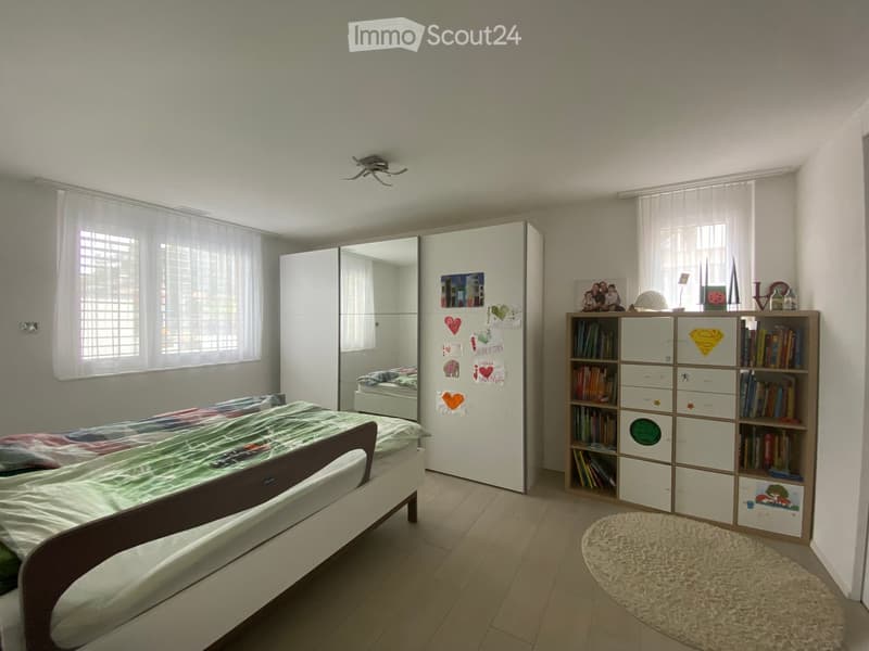 Bright bedroom with a single bed, colorful bedspread, large mirrored wardrobe, bookshelf filled with books, and a round shaggy rug on the floor.