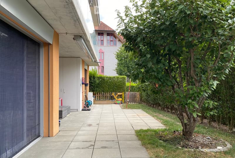 Garden area with a paved pathway, small tree, shrubs, and a fenced play area with toys.