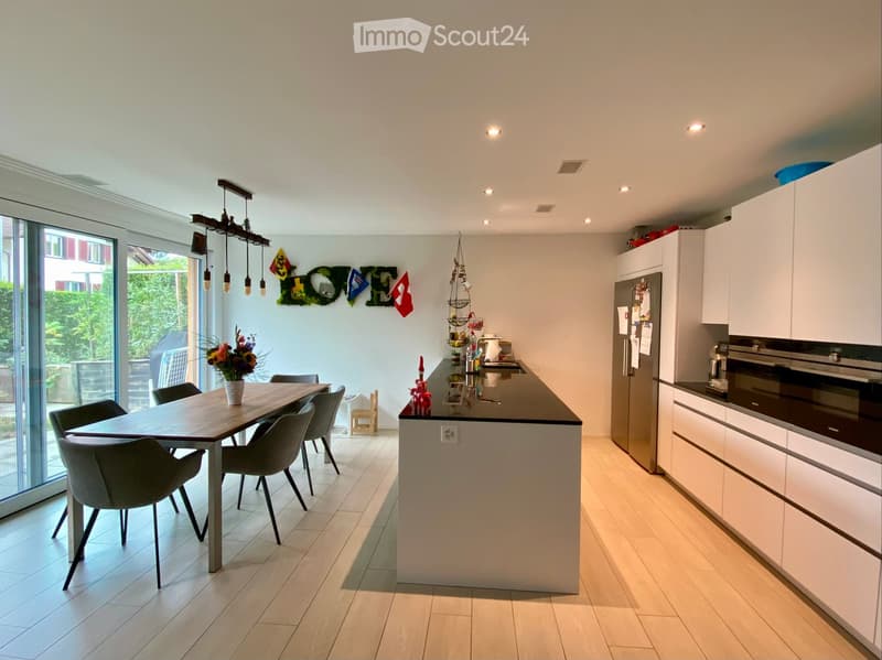 Modern kitchen with white cabinets, a central island, a dining table with chairs, and a vase of flowers. The space features recessed lighting and a pendant light above the island.