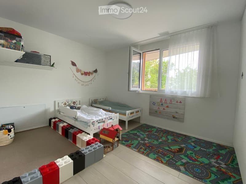 Brightly lit children's bedroom with two single beds, colorful Lego bricks, a toy car mat, and a small toy house on a stand. Shelves hold storage boxes and toys, with a decorative banner above the beds.
