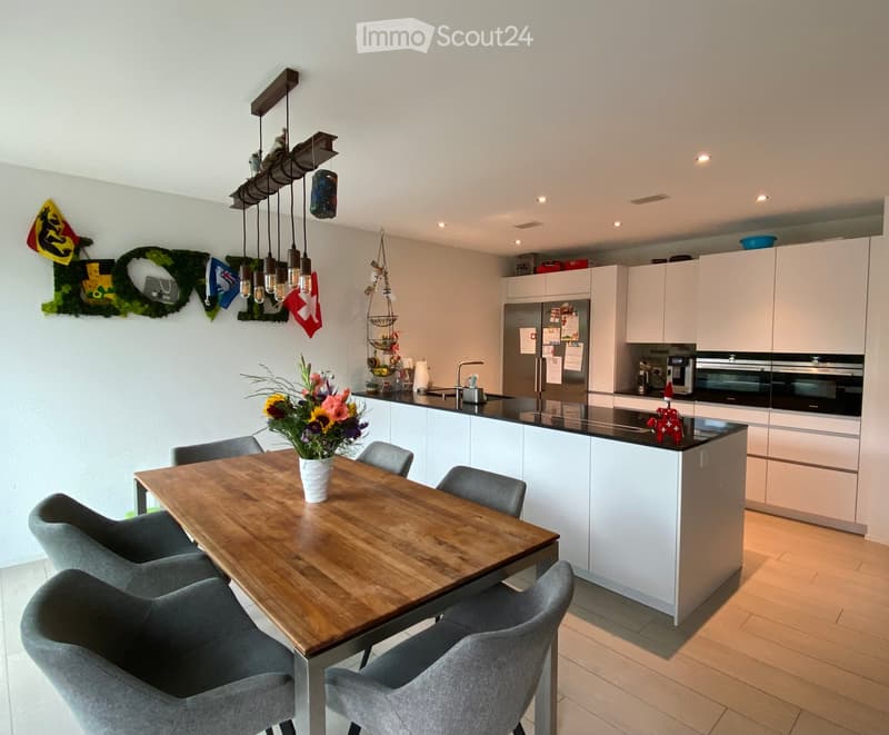 Modern kitchen with white cabinets, stainless steel appliances, a wooden dining table with six chairs, and a flower vase on the table. The kitchen features a pendant light fixture and a decorative wall with flags.