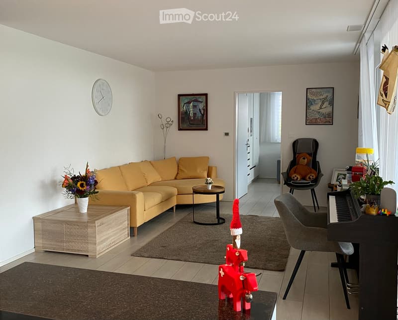 Bright living room with a yellow sofa, coffee table, side table, piano, and decorative items including a teddy bear and a small red elf figure.