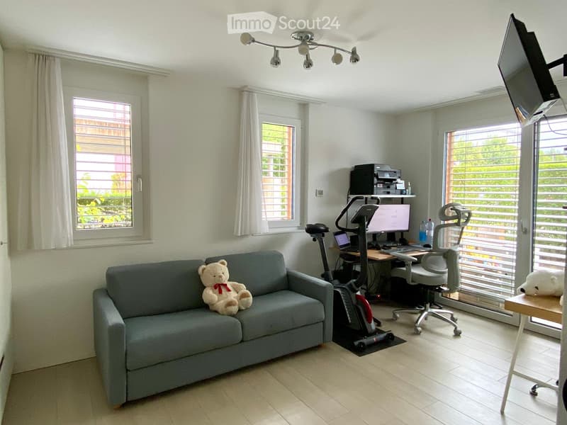 A multi-functional room with a sofa, exercise bike, desk, and computer setup. The room features large windows with blinds and a modern lighting fixture.