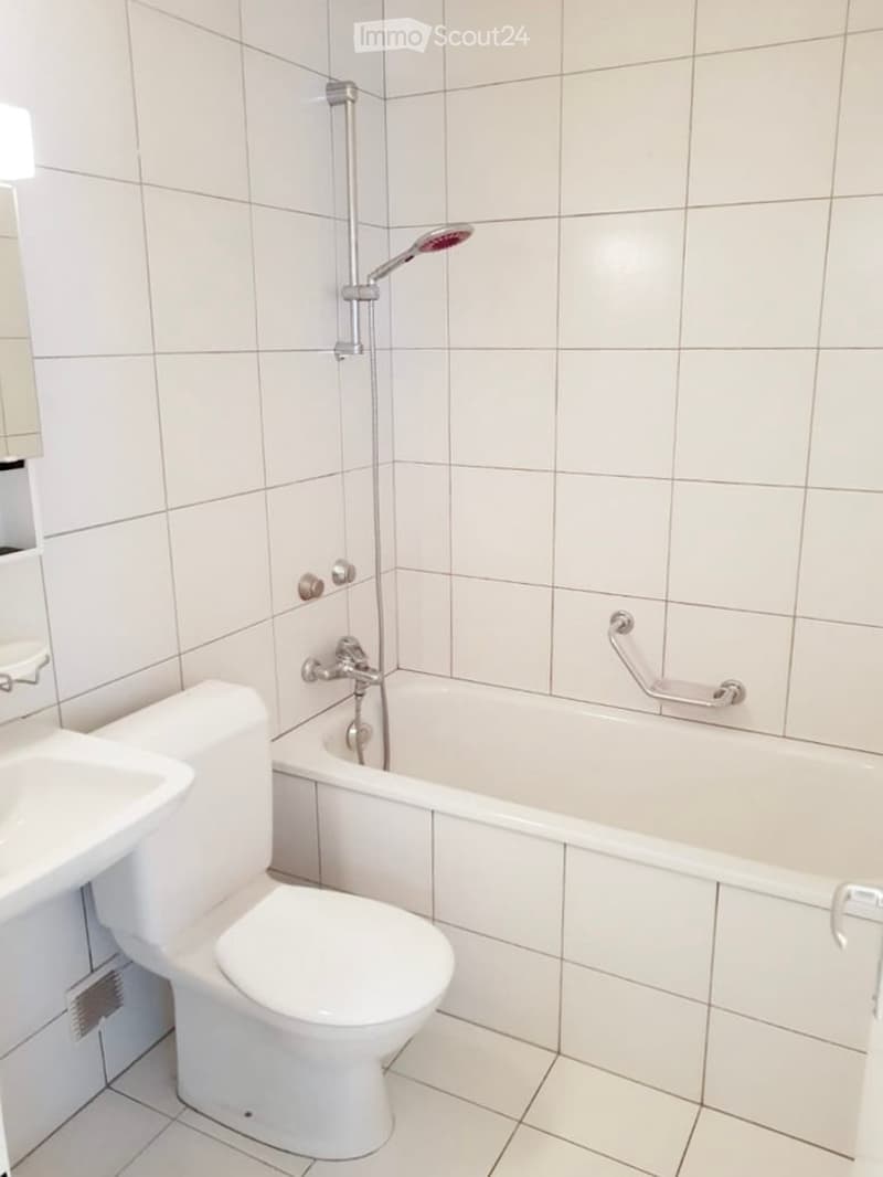 Modern bathroom with white tiled walls and floor, featuring a toilet, bathtub, sink, and a handheld showerhead.