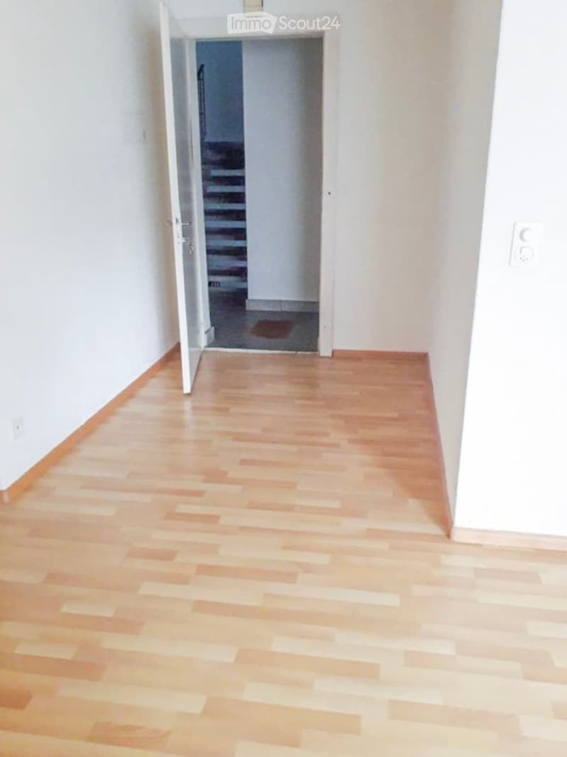 Empty hallway with light wooden flooring and white walls, leading to an open doorway that reveals a staircase.