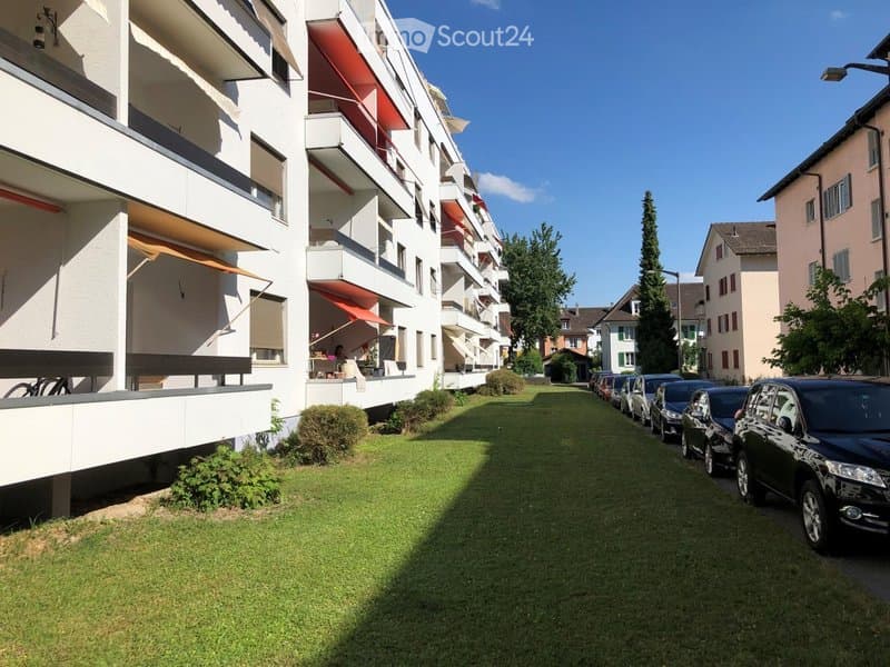 Modern multi-story apartment building with balconies, red awnings, and a well-maintained lawn in front. Several parked cars line the street.