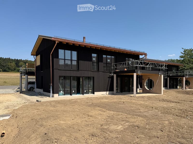 Modern two-story building with dark wooden facade, large windows, and a raised ground floor. Features a balcony and a covered entrance area.