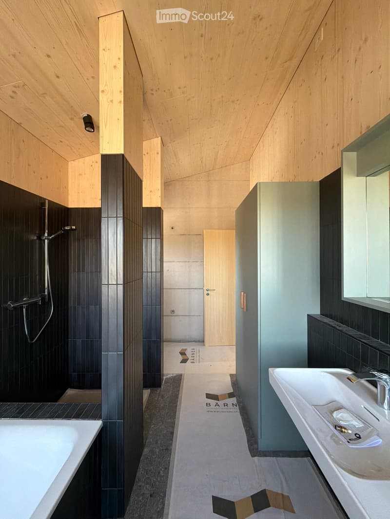 Modern bathroom with a bathtub, sink, and shower area. Features wooden paneling on the walls and ceiling, black tiles in the shower area, and a sleek, minimalist design.