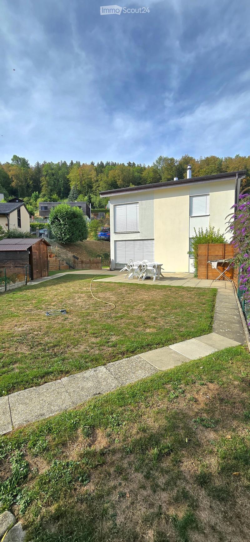 A well-maintained garden area with a small patio, outdoor furniture, and a wooden shed, surrounded by greenery and neighboring houses.