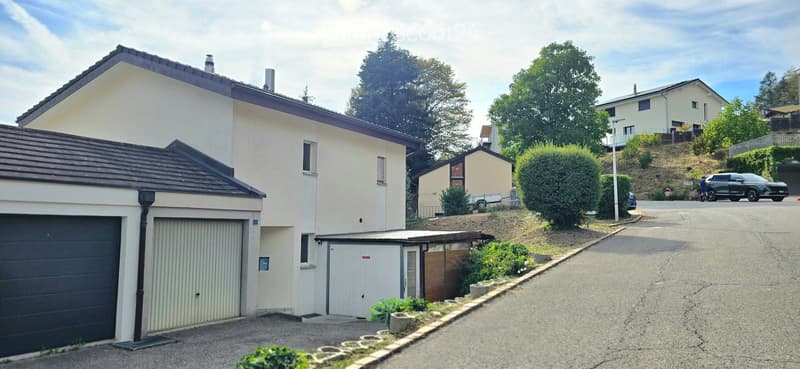 Two-story house with a garage, light-colored exterior, and a small garden area. The house is situated on a sloped street with neighboring houses and trees visible in the background.