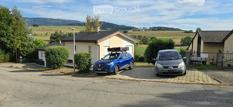 Two-story house with beige walls and dark roof, surrounded by greenery and parked cars.