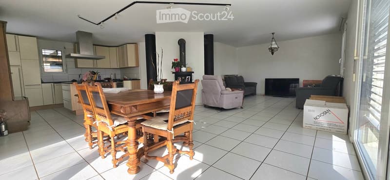 Open-plan living and dining area with tiled flooring, a wooden dining table with four chairs, a grey sofa, a television, and a modern kitchen with cabinets and appliances.