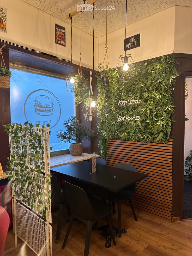 Modern restaurant interior with wooden floors, black dining table, chairs, hanging plants, and decorative signs.