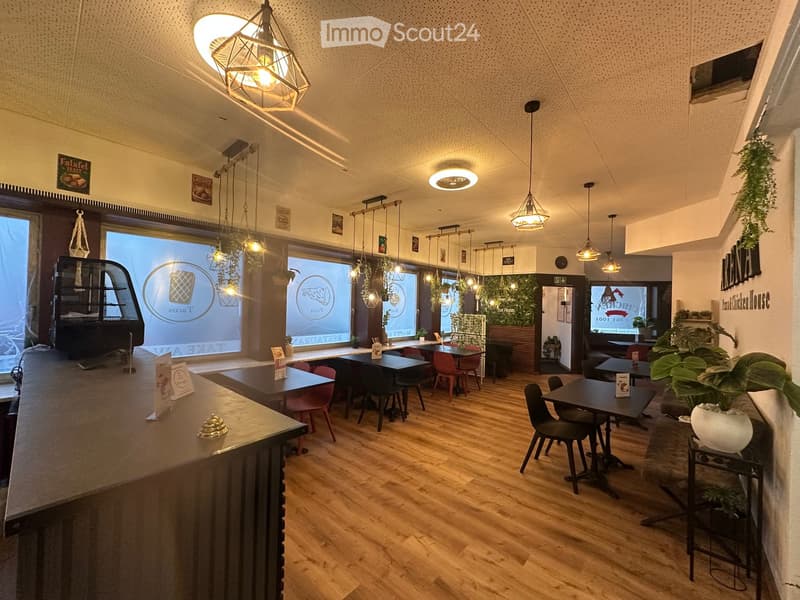 Modern restaurant interior with wooden floors, pendant lights, tables, chairs, and a counter. Large windows and plants add to the ambiance.