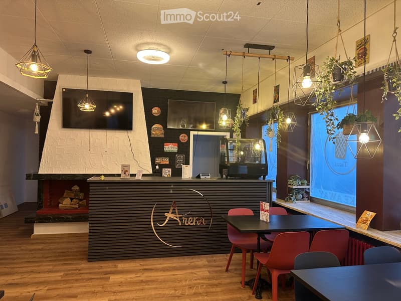 Modern restaurant interior with a reception counter, seating area, pendant lights, and a fireplace. Features include a TV screen, menu boards, and hanging plants.