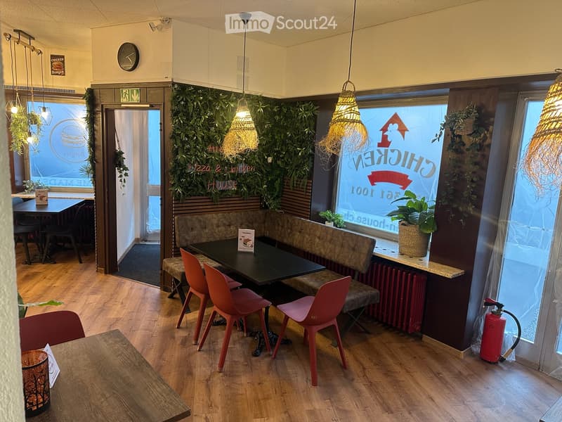 Modern restaurant interior with wooden flooring, tables, chairs, a bench seating area, hanging pendant lights, and a green wall with signage.