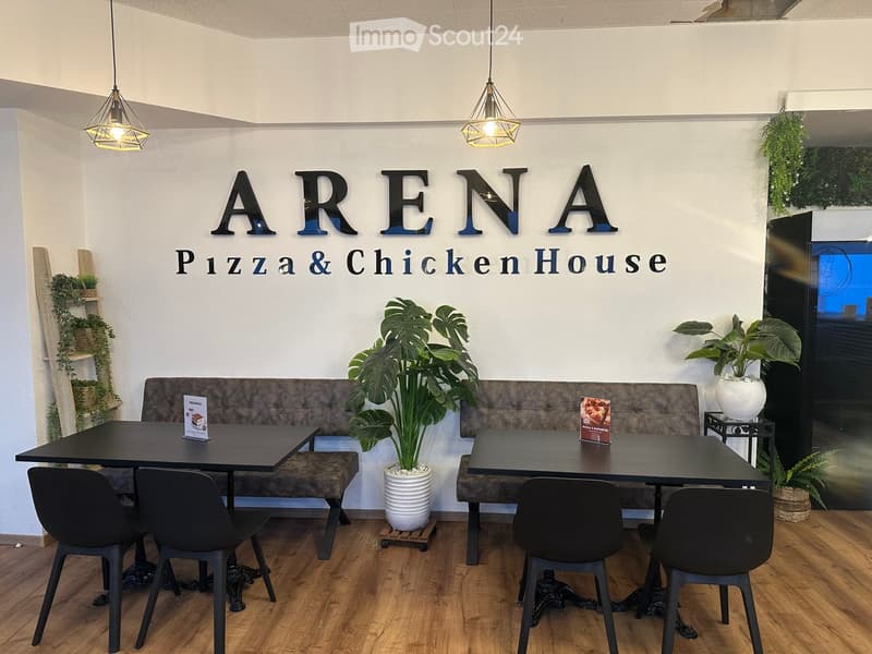 Modern restaurant interior with wooden floors, pendant lights, a long bench seating area, tables, chairs, and decorative plants.