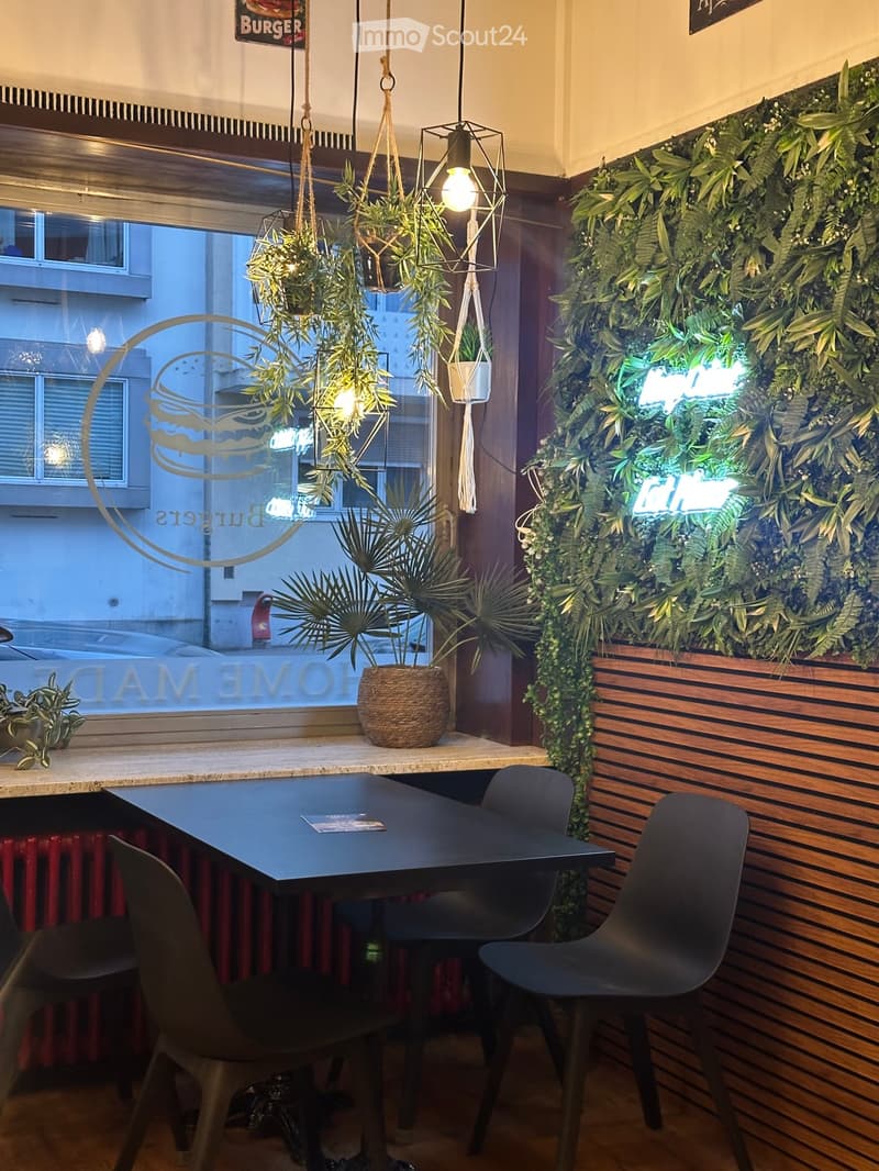 Modern interior with a black table and chairs, hanging plants, and a large green wall decoration. The space features a wooden floor and a window view of the street.