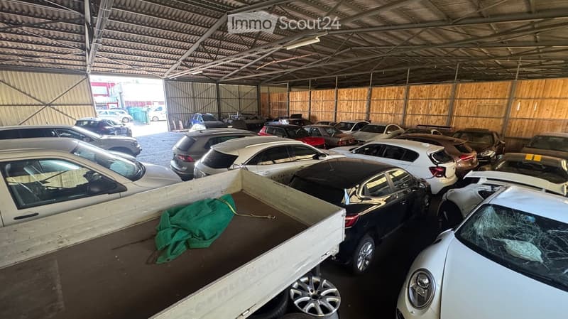 Large indoor parking area with numerous cars parked inside a metal-roofed structure. The space includes a truck bed with a green tarp, and the area is well-lit with natural light coming from the open doorway.