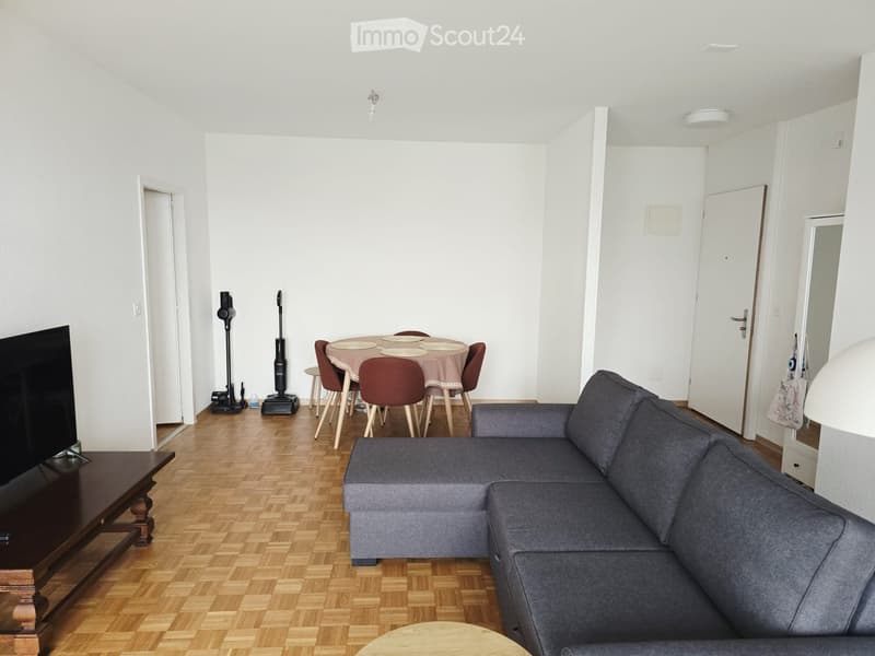Living room with a grey sectional sofa, coffee table, dining table with chairs, and vacuum cleaners in the corner.