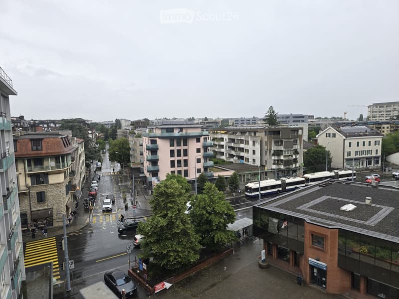 A city street view with wet pavement, multiple buildings, parked cars, buses, and pedestrians. The buildings vary in architectural style, with some featuring balconies and others having flat roofs.