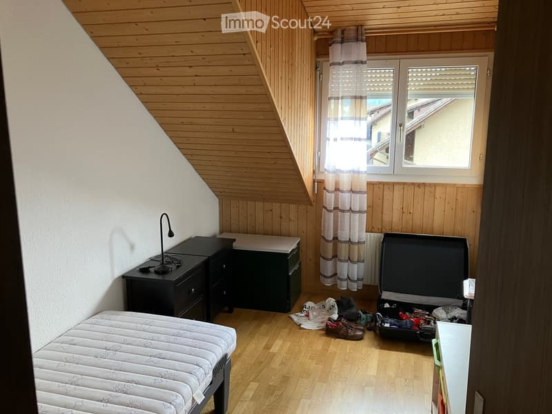 Cozy attic bedroom with wooden walls and ceiling, featuring a single bed, nightstand with lamp, open suitcase, and scattered items on the floor.