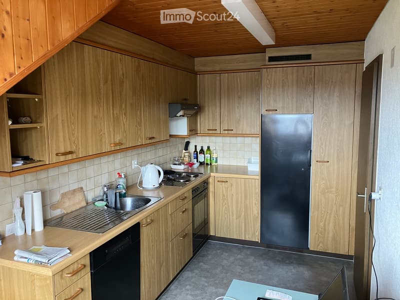 The kitchen features light wooden cabinets, a stainless steel refrigerator, a sink with a faucet, a stove, and a dishwasher. The countertops are light-colored, and there are various kitchen items such as a kettle, bottles, and a cutting board.