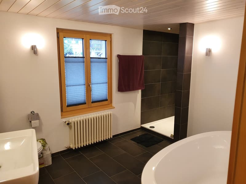 Modern bathroom with a bathtub, sink, toilet, radiator, and a towel hanging on the wall. The room features dark tiled flooring and a large window with blinds.