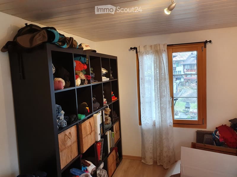 A room with a large black shelving unit filled with books, clothing, and other items. A window with a white curtain offers a view of the outdoor area. The floor is wooden, and there are additional items scattered around the room.