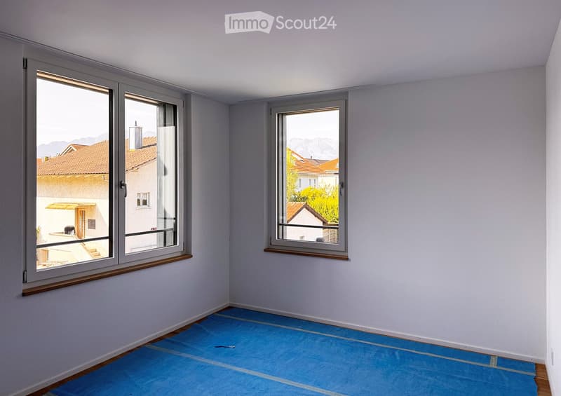 Empty room with light grey walls, blue protective floor covering, and large windows offering views of neighboring buildings and mountains.