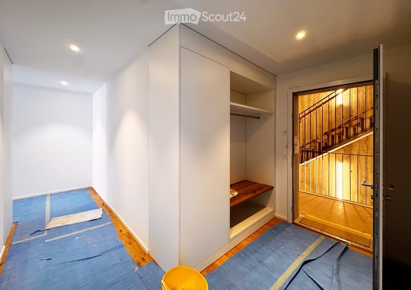 Empty hallway with white walls, wooden flooring, and a built-in closet with shelves. A doorway leads to a staircase with wooden steps and a metal railing.