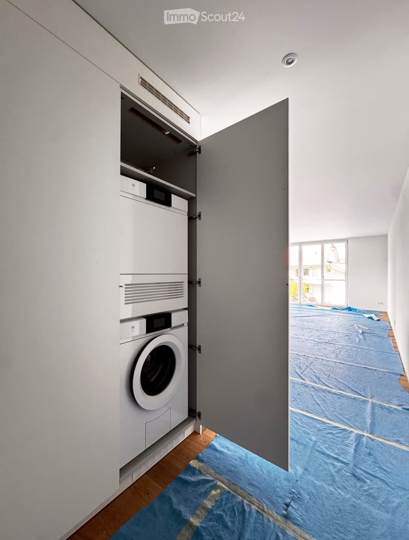 Empty room with wooden flooring and a blue protective sheet covering part of the floor. A built-in laundry unit with a washing machine is visible on the left side.