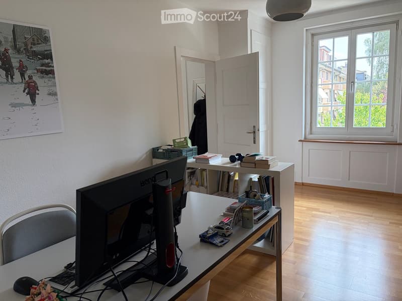A well-lit room with wooden flooring, a large window, a desk with a screen, laptop, chair, and various items including books, a cup, and a toy car. A large picture frame is on the wall, and there is a shelf with more books and items.