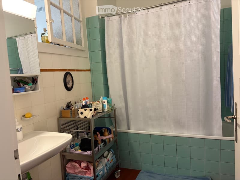 Bathroom with a sink, mirror, shower curtain, and a cart holding various toiletries and items. The walls are tiled in white and green, and there is a framed picture on the wall.