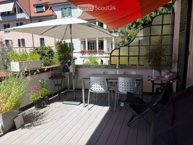 A modern wooden terrace with a dining set, umbrella, and potted plants, overlooking a cityscape with neighboring buildings.