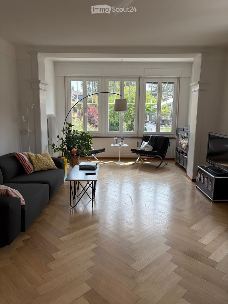 Modern living room with wooden herringbone flooring, large windows, a dark grey sofa with patterned cushions, a black coffee table, a white side table, a floor lamp, a TV stand, and a potted plant.
