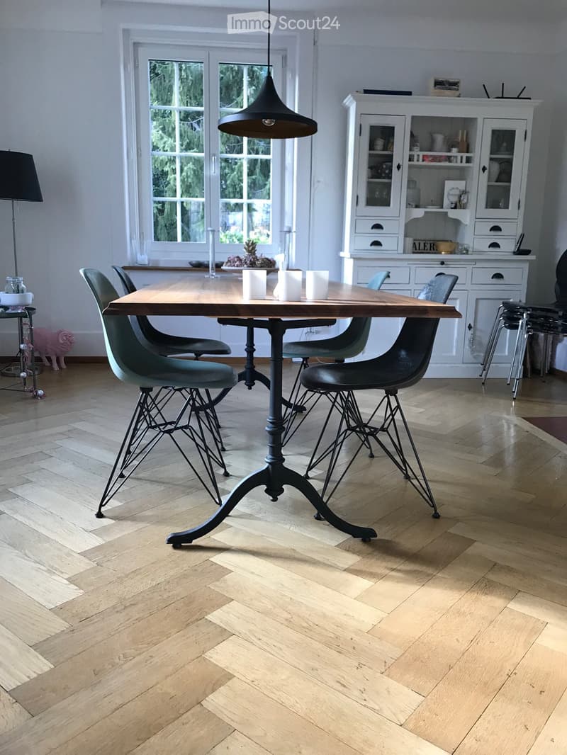 Modern kitchen with herringbone wooden flooring, a large wooden dining table with four chairs, white cabinets, and decorative items on the counter.