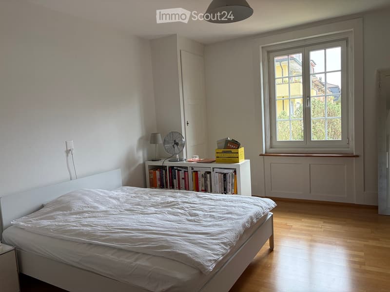 Bright bedroom with wooden flooring, a white bed, a bookshelf filled with books, a small fan, and a table lamp. Large window with view of greenery.
