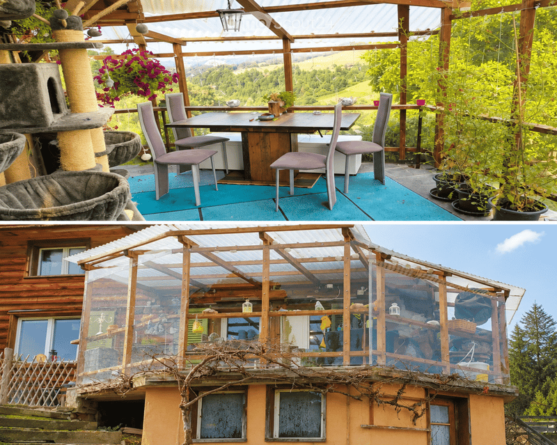 A spacious, open-air balcony with a wooden frame, glass panels, and a scenic view of green hills. Features a dining table with chairs, a cat tree, and potted plants.