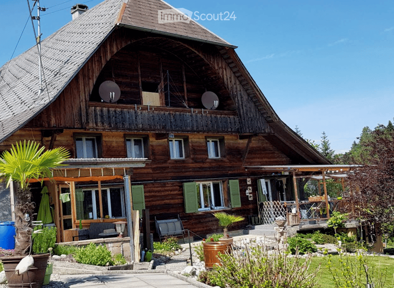 A two-story wooden house with a distinctive curved roof, multiple windows, and a balcony. The house features a well-maintained garden with potted plants and outdoor seating.