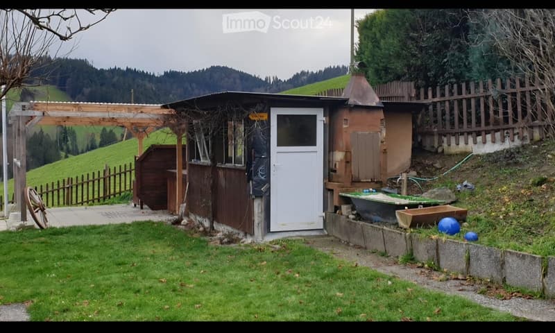 A rustic outdoor area with a wooden pergola, small shed, fenced yard, and a view of green hills and trees in the background.